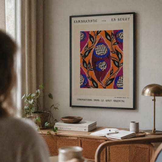 A framed art print featuring a vibrant, colorful pomegranate pattern in shades of orange, purple, blue, and green, styled on a beige wall in a cozy, sunlit room with a wooden desk, books, and a brass lamp.
