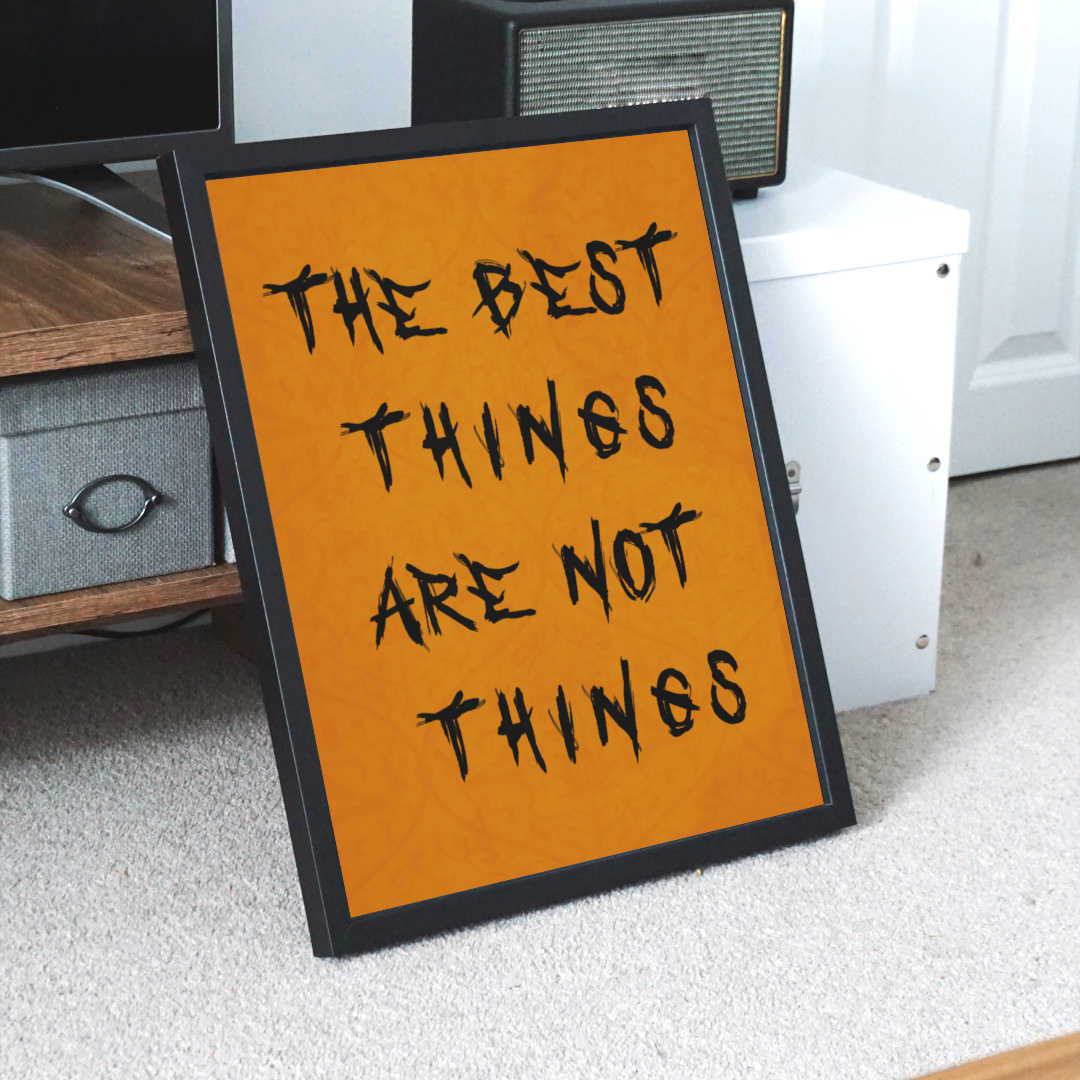 Framed motivational wall art poster with the quote "The Best Things Are Not Things" in bold black font on an orange background, leaning against furniture in a modern home interior setting—ideal for inspirational home or office decor.