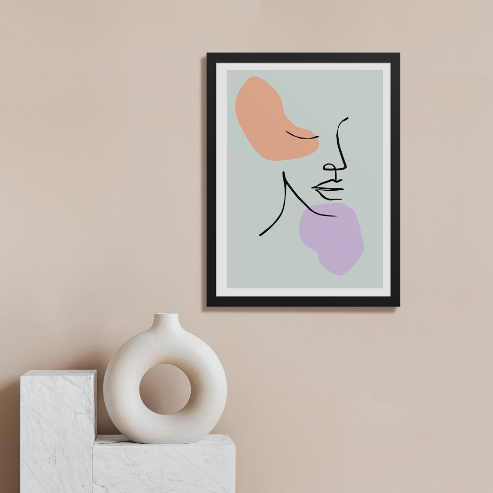 A contemporary interior with a beige wall displaying the same black framed abstract line art print containing peach and lavender shapes with curved lines. On a surface below sits a white circular sculptural vase or decorative object with a hollow center.