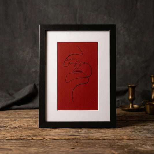 A framed minimalist art print featuring a black continuous line drawing of a face on a textured red background, set in a black frame with a white mat, displayed on a rustic wooden surface against a dark gray backdrop.
