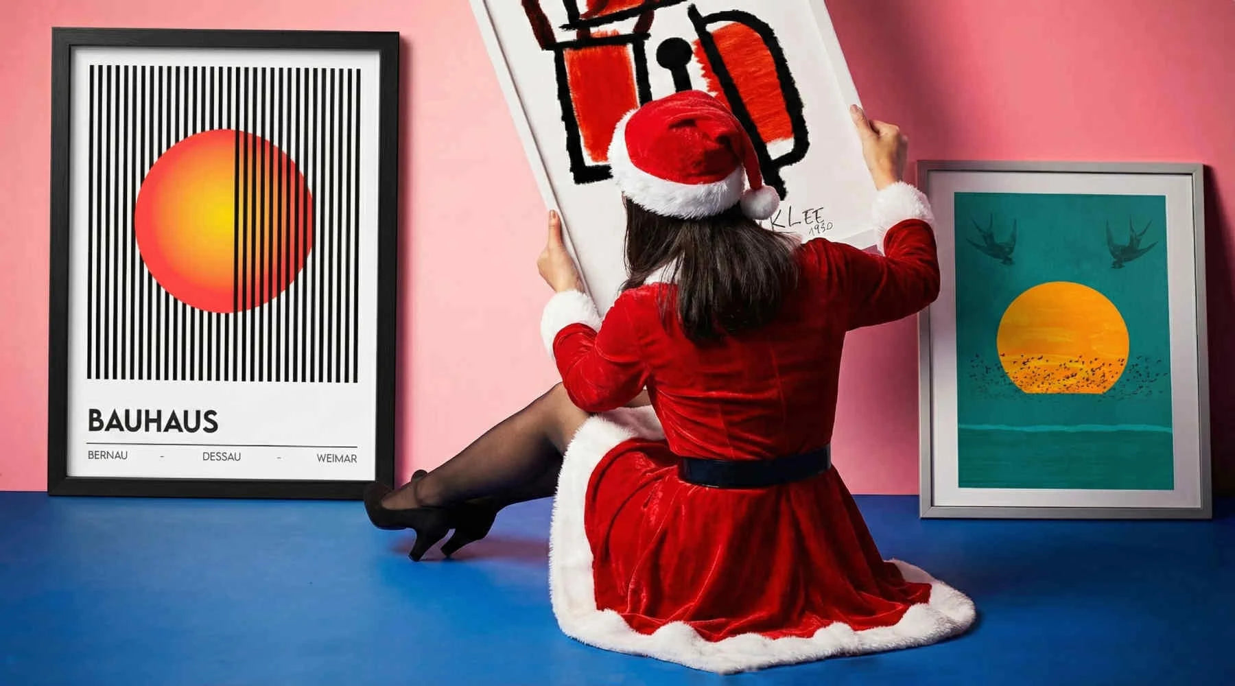 Person in a red dress and Santa hat sitting on a chair with art prints on a pink wall.