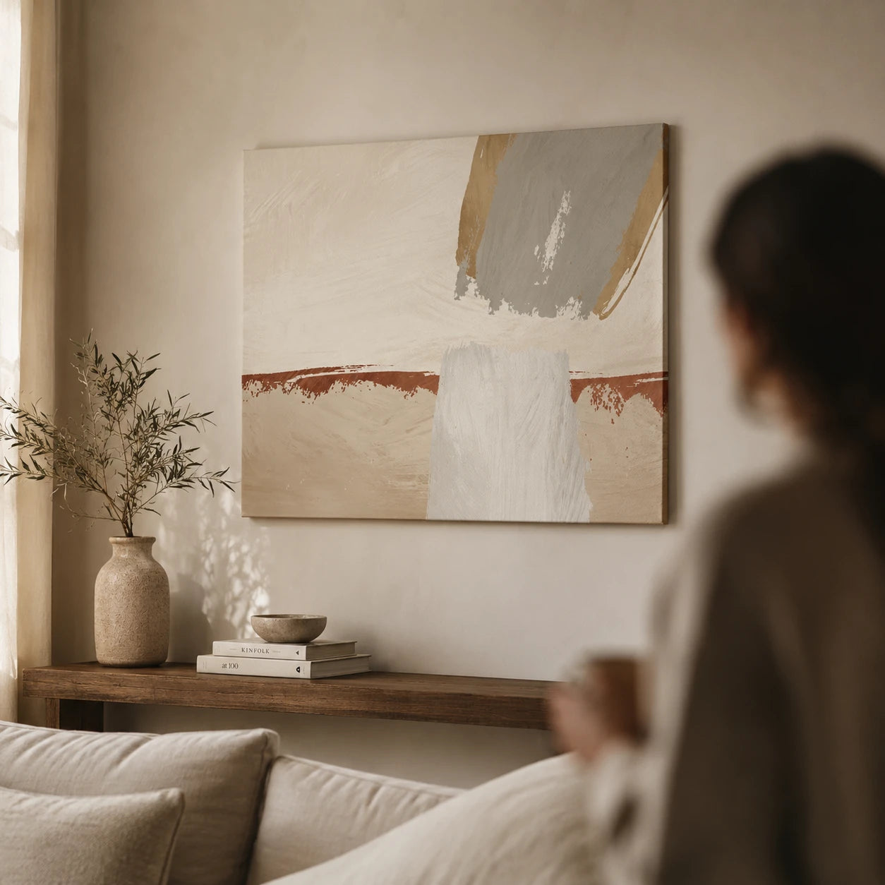 An abstract canvas painting with beige, white, gray, and terracotta brushstrokes hangs on a warm beige wall in a softly lit living room. A person is blurred in the foreground, seated near a sofa and a wooden console table with a vase and books.