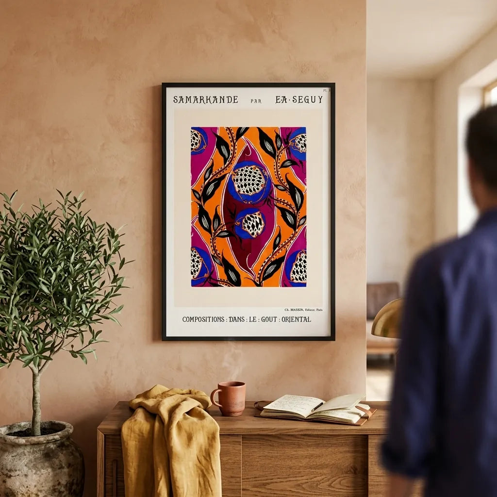 A framed art print featuring a colorful, patterned design with French text at the top and bottom, hanging on a textured beige wall in a styled room. A person is visible in the blurred background on the right, and a potted plant and wooden console table are in the foreground.