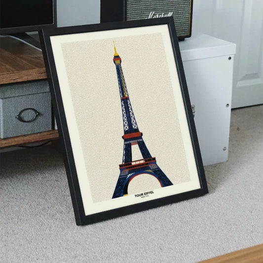 Minimalist Bauhaus Eiffel Tower print in a black frame, with the Paris landmark in navy, red and yellow geometric detail on a textured cream background, shown leaning on a floor in a living room.