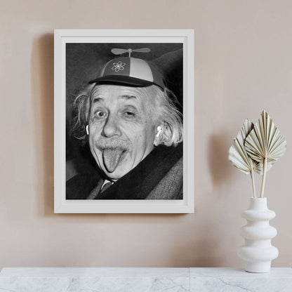 Black and white portrait photograph of Albert Einstein sticking his tongue out, wearing a propeller hat decorated with an atom symbol, vintage poster print. On beige wall.