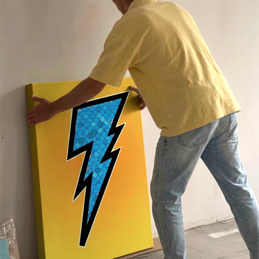 A man in a yellow short sleeved polo shirt and light blue jeans holds a large yellow canvas print featuring a blue lightning bolt with a stained glass pattern against a neutral wall in a softly lit room.